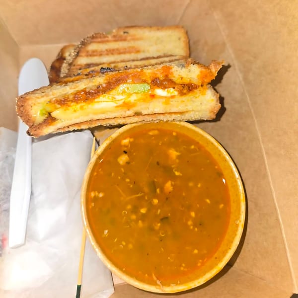 Grilled cheese sandwich with a side of soup, packaged in a brown takeout box.